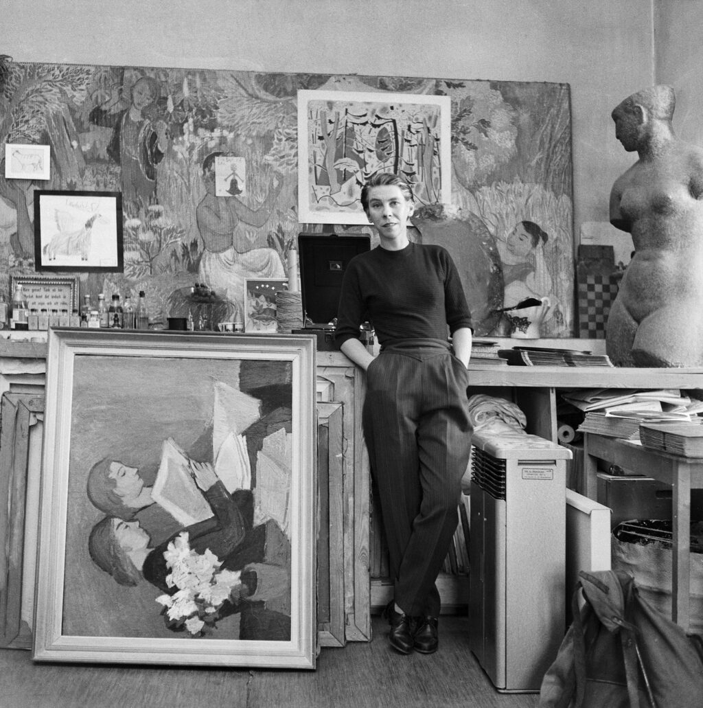 Mustavalkoinen valokuva, jossa nuori nainen seisoo ateljeessa maalaukseen nojaten.