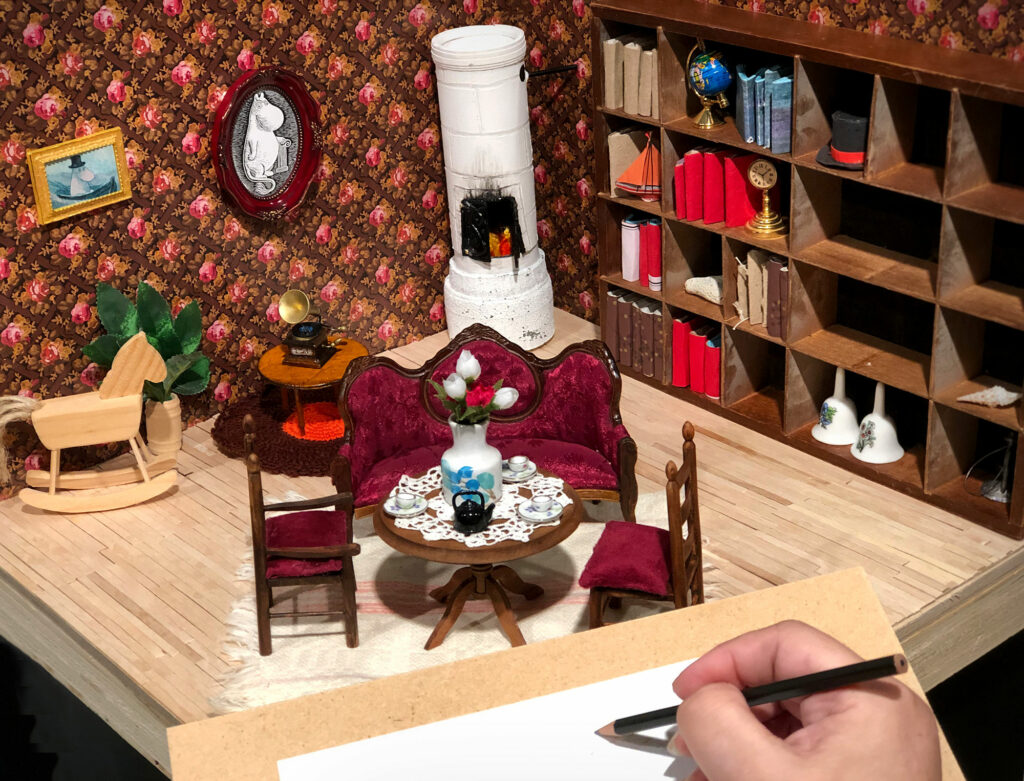 Kuva miniatyyrihuoneesta, jonka etualalla näkyy piirtävä käsi.